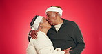 Christmas, kiss and face of couple in studio for love, holiday season and festive celebration. Romance, xmas vacation and bonding with elderly man and woman on red background for tradition and smile