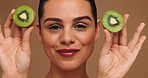 Face, woman and kiwi for skincare in studio for vitamin c,  anti aging benefits and hydration. Smile, person and bite fruit for beauty, organic ingredients and natural facial glow on brown background