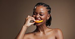 Beauty, face and orange with black woman in studio for detox, diet or health eating benefits. Biting, fresh and fruit with happy African model on brown background for organic nutrition or vitamins