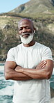 Face, happy man and fitness at beach with arms crossed, exercise goals and earphones for music motivation. Mature, black person and smile at sea with audio tech for radio, workout recovery and break.