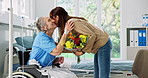 Senior mother, daughter and hug in hospital with flowers for comfort, talking or bonding together. Elderly mom, mature woman and discussion in wheelchair with bouquet, embrace and get well soon wish.