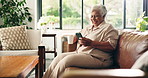 Coffee, mobile and senior woman in home for relax, news or laughing at meme in retirement on sofa. Tea, smile and elderly person with phone for joke, online communication or reading funny article