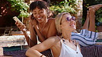 Music, phone and women at swimming pool together for audio, song or sound streaming in summer. Earphones, relax and smile with friends sunbathing outdoor on holiday for travel break, trip or vacation