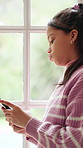 Sad, child and typing in home with phone, cyberbullying and shame for gossip on social media post. Window, unhappy kid or girl with depression, mental health or disappointed for online rumor in house