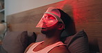 Red light, relax and man with mask for skincare, beauty or wellness with collagen production. Health, cosmetic and male person on bed with facial glow therapy for dermatology treatment in home.