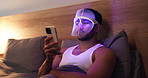 Blue light, mask and man with phone for skincare, relax or wellness with antibacterial facial. Health, beauty tech and male person on bed with LED phototherapy for dermatology treatment in home