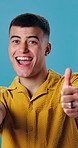 Thumbs up, excited and face of man in studio with positive review, good news and yes sign. Smile, emoji and portrait of person with gesture for great feedback, approval or vote by blue background.