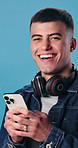 Happy man, phone and face in studio for social media, funny joke or meme on app. Male student, portrait and smile on tech for networking, chatting or headphones or communication on blue background