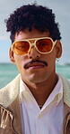 Face, style and sunglasses with man on beach for holiday break, travel or vacation in summer. Retro fashion, shades and trip with serious person outdoor in nature for tourism or weekend getaway